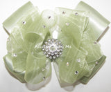A large light sage green organza hair bow with a silver rhinestone in the center and small scattered rhinestones attached to a choice of a clip or headband